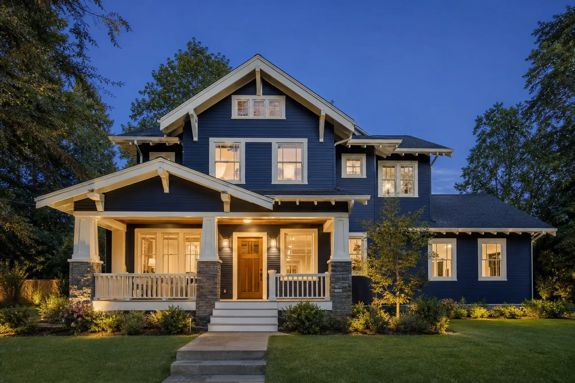 Navy Craftsman house with crisp white trim, a natural wood entry door, and warm porch lights at twilight.