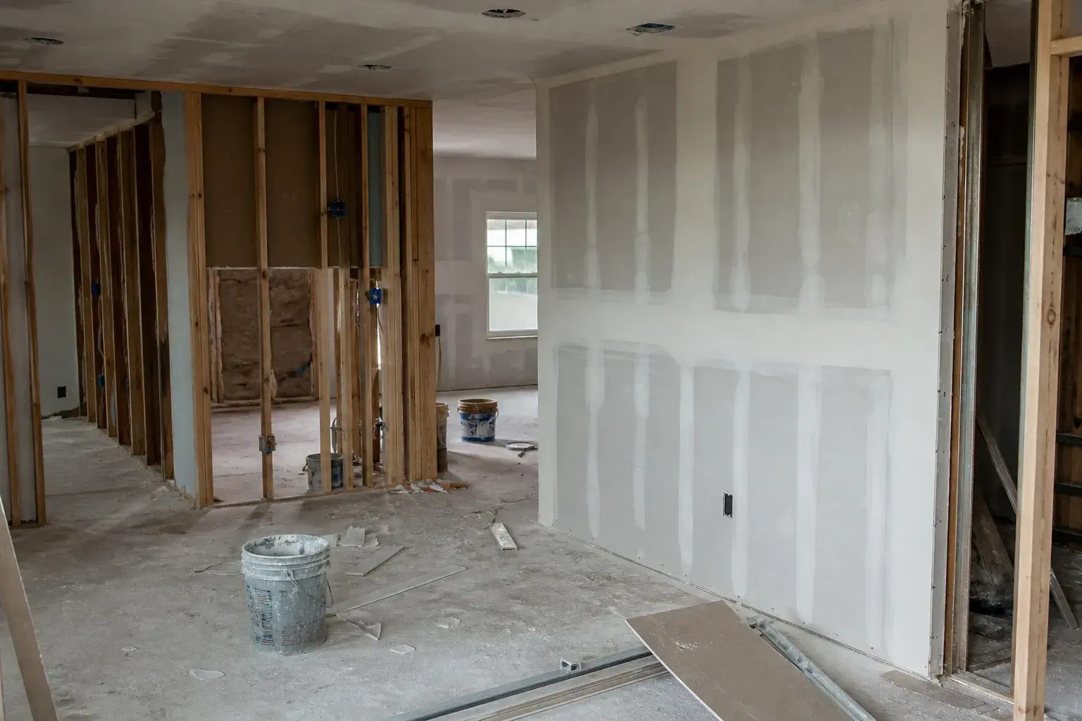 Drywall mid-job: one wall hung, taped, and mudded, the adjoining walls still open studs with insulation, mud buckets on the slab.