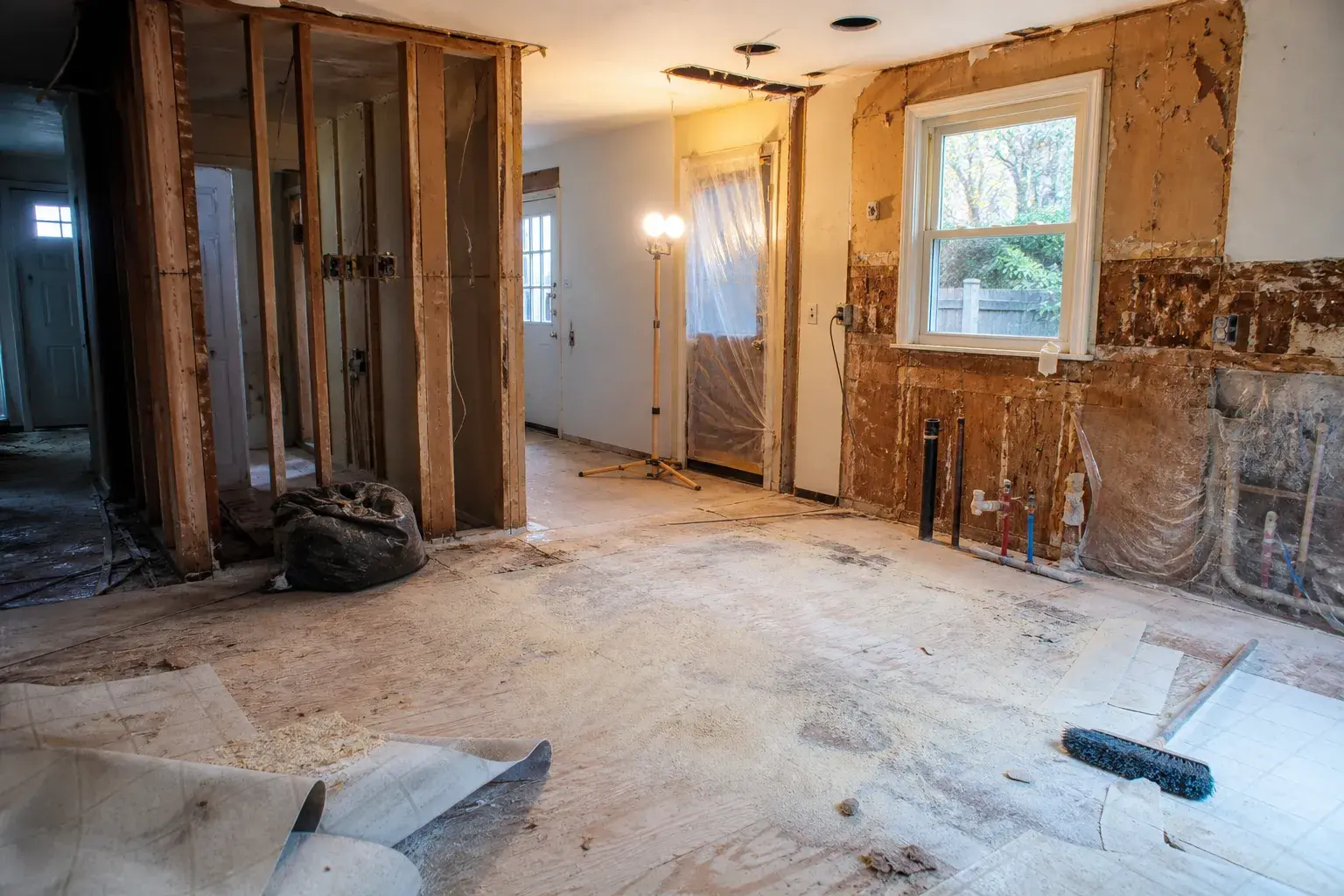 Kitchen after demo: walls open to the studs, old plumbing stubs exposed on the window wall, a work light on a tripod, and the door sealed in plastic.